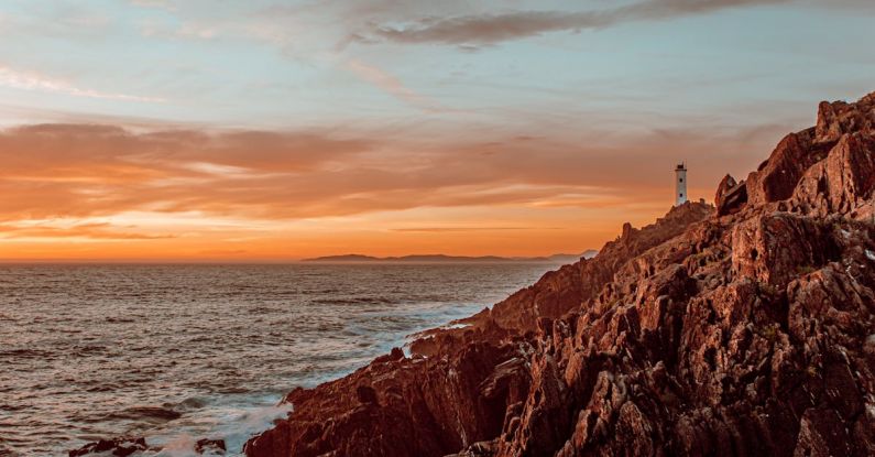 Stone Setting - Amazing scenery of lighthouse in distance located near rocky coast with waving ocean with sun setting on horizon in summer evening