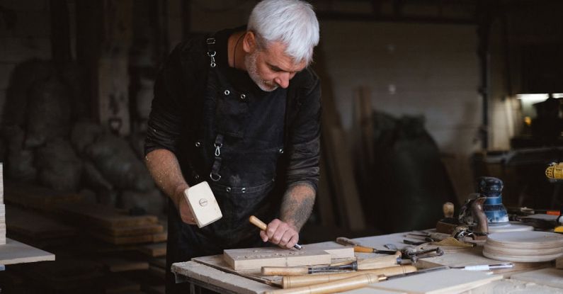 Hammering Technique - Serious male woodworker in apron using mallet and chisel to carve wooden board at table with abundance of instruments in carpentry