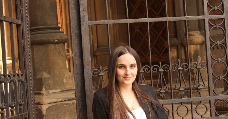 Iron Gates - Young Brunette Woman in Blue Jeans and Black Jacket Sitting at Church Gates