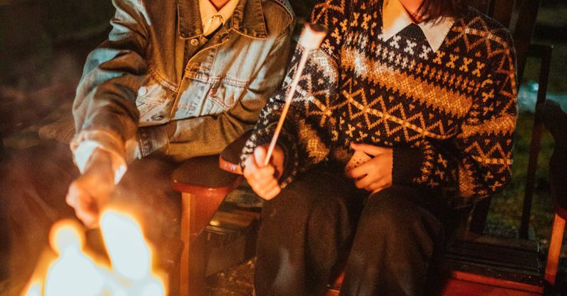 Heat Treating - Smiling teen girlfriends in warm clothes sitting in wooden armchairs near bonfire in backyard while frying marshmallow at night